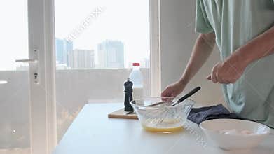 Man seasoning and whisking beaten eggs in glass bowl with pepper grinder in kitchen with milk during breakfast