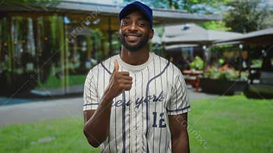 Man wearing baseball jersey and blue cap standing, looking to the side in front of a building; relaxed casual leisure