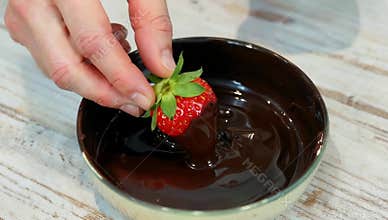 Hand dips fresh strawberry into melted dark chocolate in a bowl on a wooden table surface