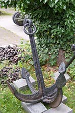 Heavy metal anchor rests on stones near chains in a green park area surrounded by trees