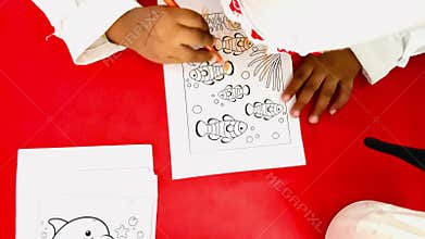 A young child sits at a red table, coloring a paper with illustrations of clownfish (Amphiprioninae)