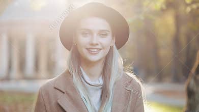 Amazing girl in coat with black hat standing down the park alley, looking into the camera and gives amazing smile. Park