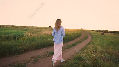 Magnificent landscape, green field, bright light sky, slender girl with blond hair walks along the road in long loose