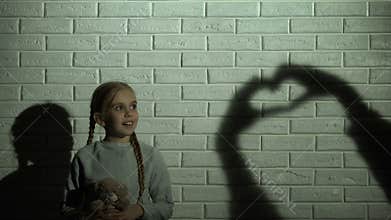 Girl looking at heart shadow on wall and hugging teddy bear, hope and kindness