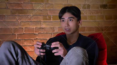 Closeup portrait of young korean man playing video cames using console sitting in the bean bag indoors