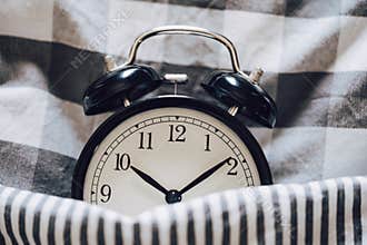 Black retro alarm clock sleeping on pillow with blanket metaphor of insomnia, late at work, well sleep with time countdown or wake