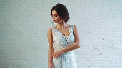 A lovely lady with dark hair in a gentle hairstyle is standing in the studio in a long elegant white dress, posing