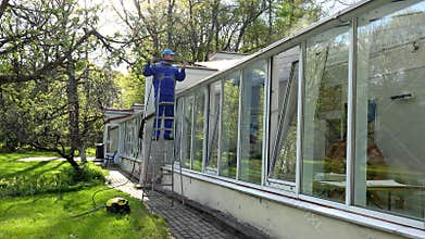 Professional worker on ladder washing glasshouse roof with high pressure tool.