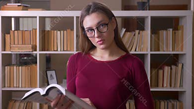 Young and beautiful brunette female student in eye glasses reading book watches into camera in library.