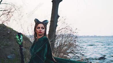 Horned femme fatale with magical green staff in hand turns back with sad face, girl in long emerald velor mantle with