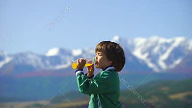 A very cute boy drinks orange juice on a background of beautiful mountains. 4k Little boy relaxes on the top of the hill