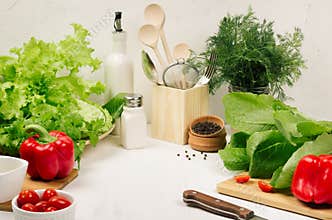 Kitchen white interior with raw fresh green salad, red cherry tomatoes, kitchenware on soft white wood table, copy space.