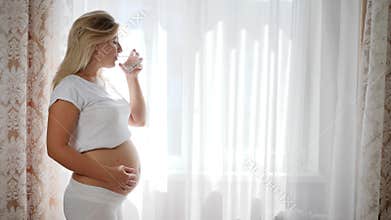 Lucky pregnant woman keeps glass pure water and rubbing big naked belly near window