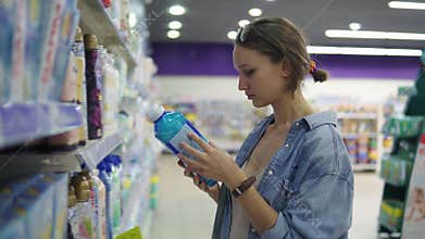 Young pretty woman in casual examines goods in a supermarket. Reads the label on the product. Selects a laundry rinse