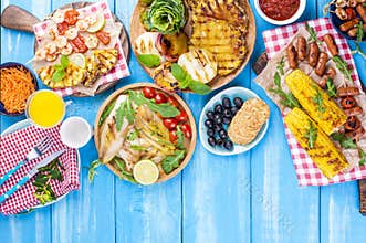 Grilled vegetables, shrimp, fruit on a wooden plate and sausages, juice and salad on a blue background. Summer dinner. Free space