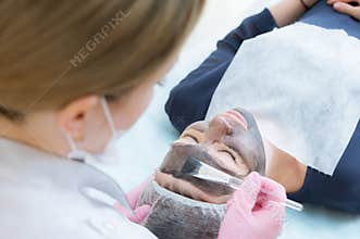 The cosmetologist in pink gloves with a brush applies a carbon mask for peeling on the face of a young girl in a