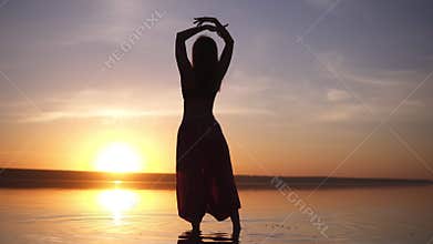 Silhouette of a slender girl walking on the water in yoga pants, raises her hands. Carefree, freedom. Incredible sunset