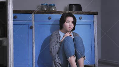 Sad lonely young Asian girl sitting on the floor in kitchen, holding her knees with arms, domestic violence concept 50