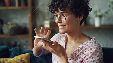 Smiling young woman is dialing telephone number then talking in loudspeaker mode holding smartphone in hand. Modern