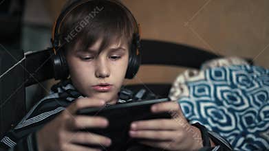 The boy blonde listens music in black headphones and plays an online game on the black phone at home