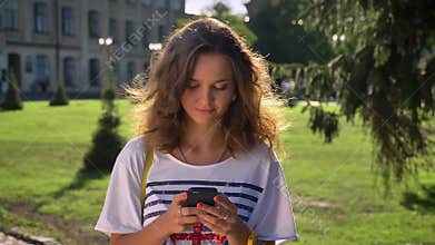 Young Caucasian girl is standing in a park and is using a smartphone, thinking, university in the background