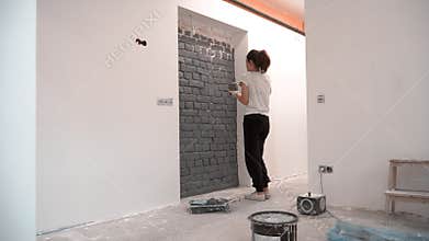 Young woman paints brick wall in gray colour using brush