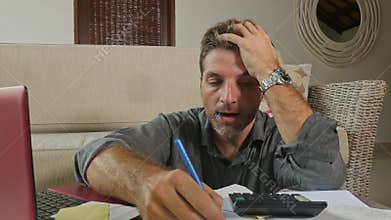 Young stressed and worried man at home living room using calculator and laptop doing domestic accounting paperwork feeling