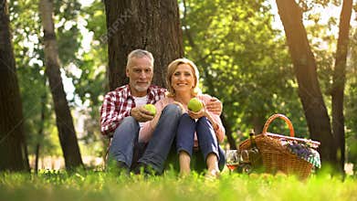 Senior couple sitting in park and eating green apples, picnic, family weekend