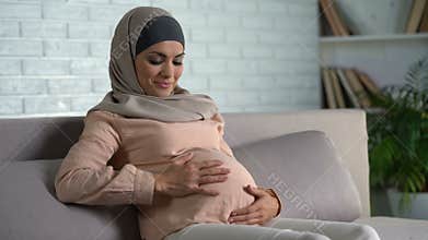 Smiling pregnant woman stroking belly, resting on home sofa, maternity happiness