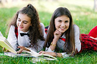 Two schoolgirls in red school uniforms are lying on a green lawn with books