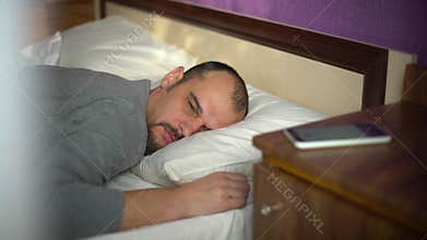 Young bearded man sleeping in bed is awaked by alarm signal on his phone