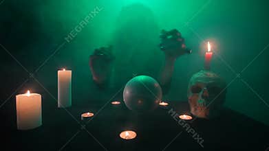 Scary witch in green smoke, conjuring on a crystal ball, occult table with a skull and candles, fast motion