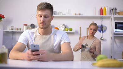 Handsome man typing message in phone, jealous wife peeking, cheating in marriage