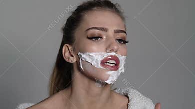 Lady shaves her face with sharp blade of straight razor. Woman with face covered with foam holds straight razor in hand