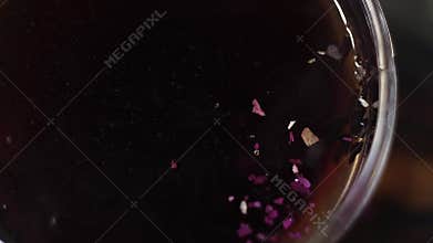 Dried purple herbs are falling on surface of hot tea liquid in glass bowl.