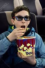 Cute young guy in a jacket eats popcorn sitting in a cinema chair. A teenager in 3D glasses puts his hand to his mouth and