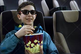 The teenager in 3D glasses smiles while watching a comedy movie. Cute European guy in a jacket sits in a cinema chair with popcorn