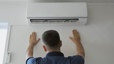 A man dripping with sweat cools under a stream of cold air standing next to a working air conditioner.