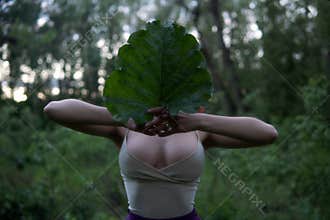 Young woman with big Tits hide her face behind a big Burdock leaf in the forest at Mysterious Twilight