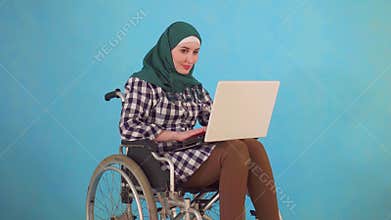 Young Muslim woman disabled in a wheelchair uses a laptop on a blue background
