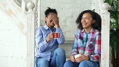 Two african american curly girls sistres sitting on stairs have fun laughing and chatting together at home