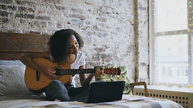 Curly african american teenager girl concentraing learning to play guitar using laptop computer sitting on bed at home