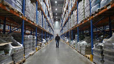 Man walking along multi level warehouse with cardboard boxes arranged on the racks to the camera