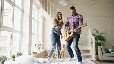 Funny happy and loving couple dance on bed singing with tv controller and playing guitar. Man and woman have fun during