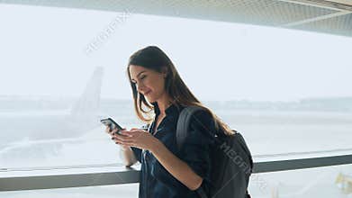 Young girl using smartphone near airport window. Happy European woman with backpack uses mobile app in terminal. 4K.