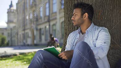 Multinational guy sitting under tree, writing in notebook, thinking, composer