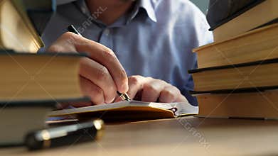 Young man writing in note on a table with piles of books. Knowledge and wisdom concept.