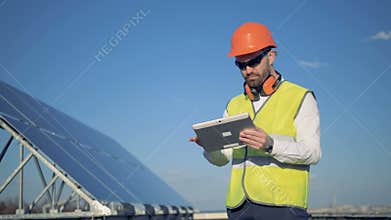 Engineers stands on a roof near solar panels, working on his gadget. 4K.