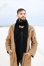 European young male person with beard wearing coat and scarf standing in white snow background.