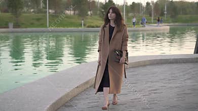 Calm young brunette in dress and coat walking near pond in autumn park